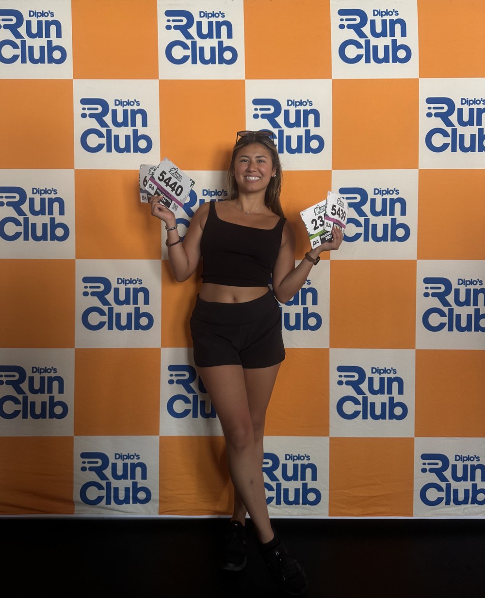 Diana Sparkles holding race bibs in front of the Diplo's Run Club backdrop